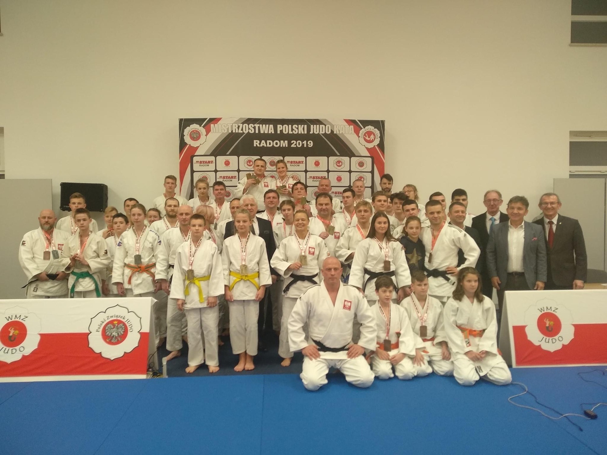 Grupa judoków z medalami na tle baneru 'Mistrzostwa Polski Judo Kata Radom 2019' oraz flagi z logo WMZ Judo i Polskiego Związku Judo.
