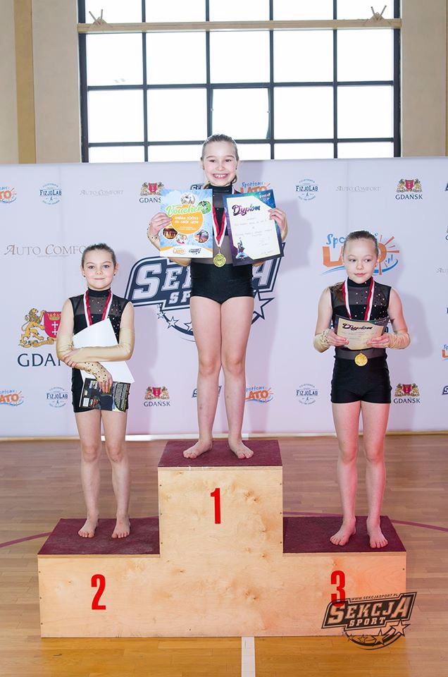 Trzy młode gimnastyczki na podium, pierwsza trzyma dyplom i voucher, druga i trzecia trzymają dyplomy, wszystkie z medalami.