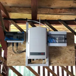 Instalacja fotowoltaiczna w drewnianej konstrukcji dachu: widoczne dwa rozdzielacze elektryczne po bokach falownika SolarEdge zamontowane na płycie OSB, kable w peszlu mocowane do belek stropowych.
