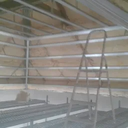 Poddasze w trakcie ocieplania, widoczne rusztowanie i drabina oparta o konstrukcję sufitu, izolacja z wełny mineralnej pomiędzy profilami metalowymi.