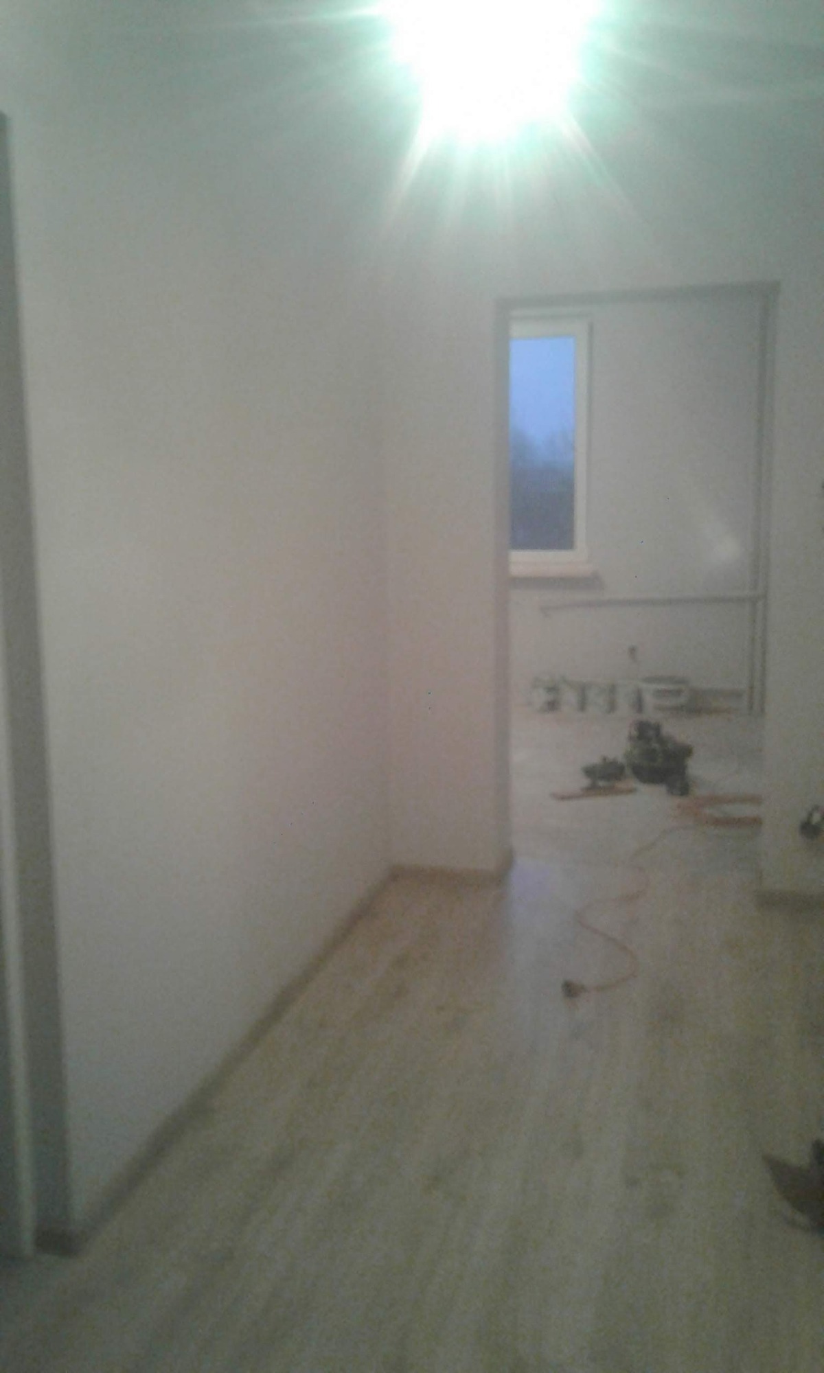 Fragment pokoju z ułożonymi panelami podłogowymi w odcieniach beżu, widoczne narzędzia i kabel elektryczny na podłodze, ściany pomalowane na biało, w tle okno z widokiem na zewnątrz.