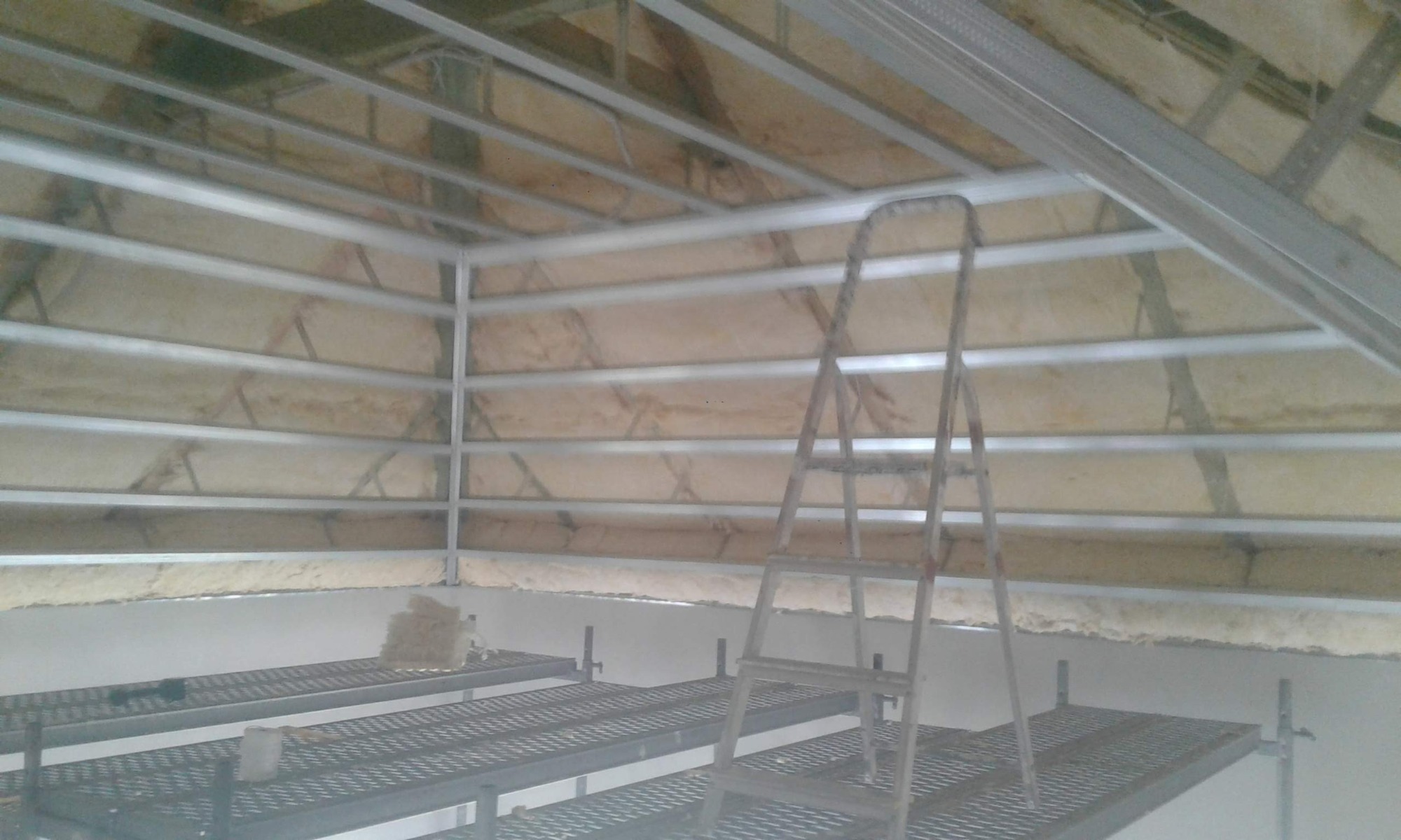 Poddasze w trakcie ocieplania, widoczne rusztowanie i drabina oparta o konstrukcję sufitu, izolacja z wełny mineralnej pomiędzy profilami metalowymi.