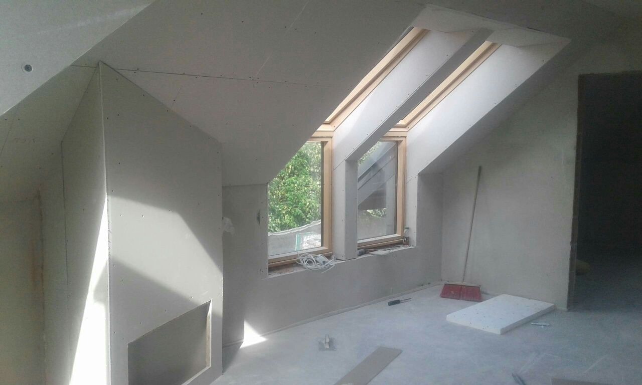 Poddasze po montażu płyt kartonowo-gipsowych, widoczne okna dachowe z drewnianą ramą, fragment ściany z otworem, narzędzia i resztki materiałów na podłodze.