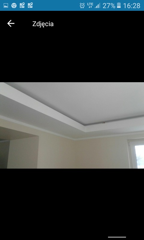 Wykończone wnętrze z białym sufitem podwieszanym z listwami dekoracyjnymi i kremowymi ścianami, widoczne okno z zewnątrz.