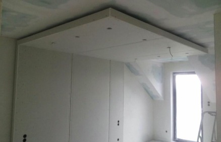 Fragment pokoju w trakcie wykańczania: zamontowana podwieszana konstrukcja z płyt gipsowo-kartonowych na suficie z przygotowanymi otworami na oświetlenie, ściany pokryte płytami GK, widoczne ślady...