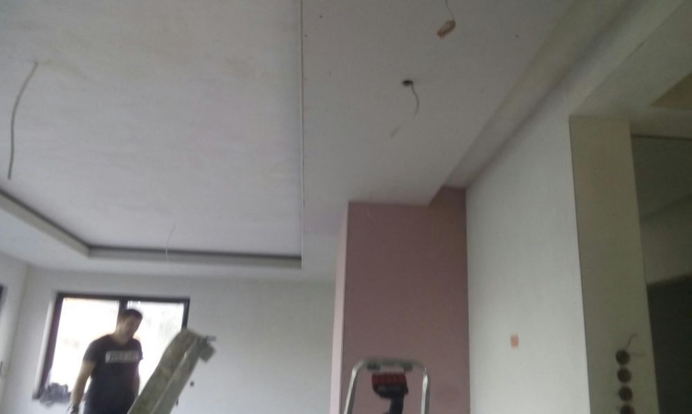 Wnętrze w trakcie wykańczania: widoczny sufit z elementami karton-gipsu, ściany przygotowane do malowania, drabina i pracownik w tle.
