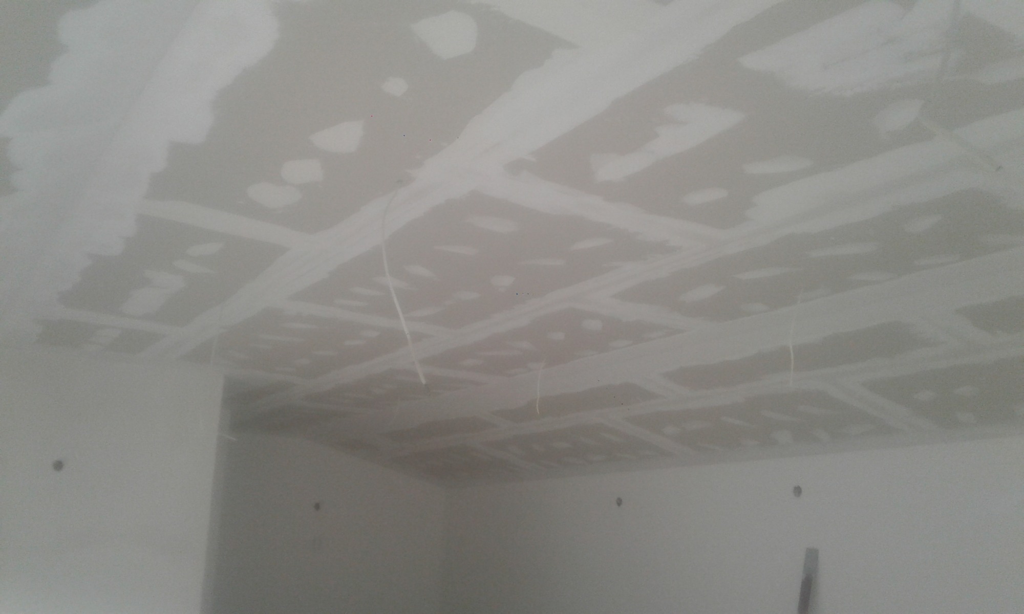 Niedokończony sufit z płyt kartonowo-gipsowych, z widocznymi łączeniami wymagającymi dalszej obróbki, w pomieszczeniu o surowych ścianach i wiszących kablach.