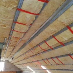 Montaż izolacji z wełny mineralnej na poddaszu, widoczne metalowe profile mocujące i czerwone taśmy zabezpieczające, surowe ściany z pustaków.