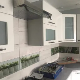 Nowoczesna kuchnia z białymi szafkami z przeszklonymi frontami, okapem i płytkami imitującymi drewno z motywem roślinnym, widoczne akcesoria instalacyjne sugerują prace wykończeniowe.