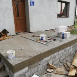 Niedokończone schody wejściowe do domu z bloków betonowych, częściowo pokryte szarą zaprawą, narzędzia budowlane na powierzchni, owczarek niemiecki leżący obok, widok na elewację budynku z oknem...