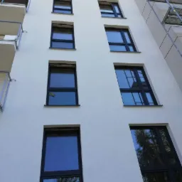 Wysoki budynek z nową, jasną elewacją i czarnymi ramami okiennymi, widziany z dołu, z fragmentem rusztowania po prawej stronie.