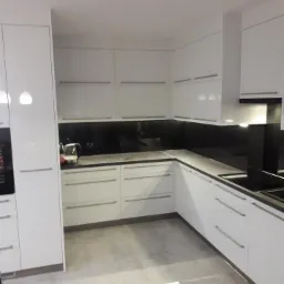 Nowoczesna kuchnia z białymi, lakierowanymi frontami, szarym blatem i czarnym szkłem na ścianie, minimalistyczne uchwyty, oświetlenie LED pod szafkami.