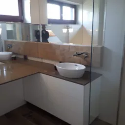 Nowoczesna łazienka z podwieszaną umywalką, brązowym blatem i lustrem z podświetlanym panelem z imitacji kamienia, widoczne drewniane panele podłogowe.