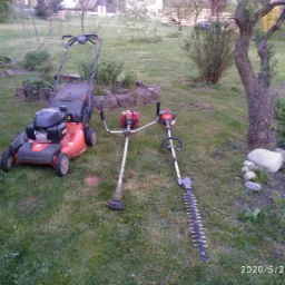 Czerwona kosiarka spalinowa, podkaszarka i nożyce do żywopłotu leżą na trawie w ogrodzie.