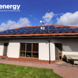 Dom jednorodzinny z panelami słonecznymi na czerwonym dachu, widoczne logo firmy '3 energy' w lewym górnym rogu, błękitne niebo z chmurami w tle.