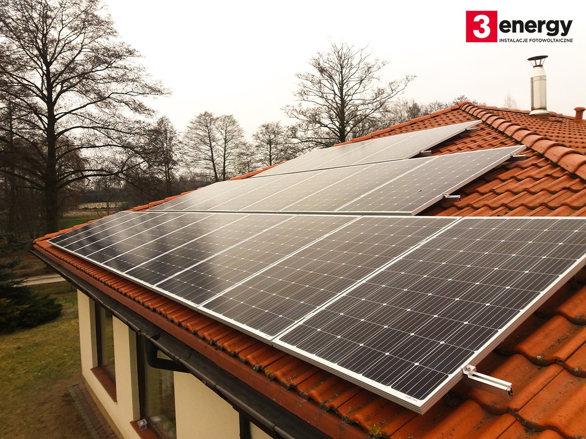 Panele słoneczne na dachu z czerwonej dachówki, widoczne drzewa bez liści w tle, logo firmy w prawym górnym rogu.