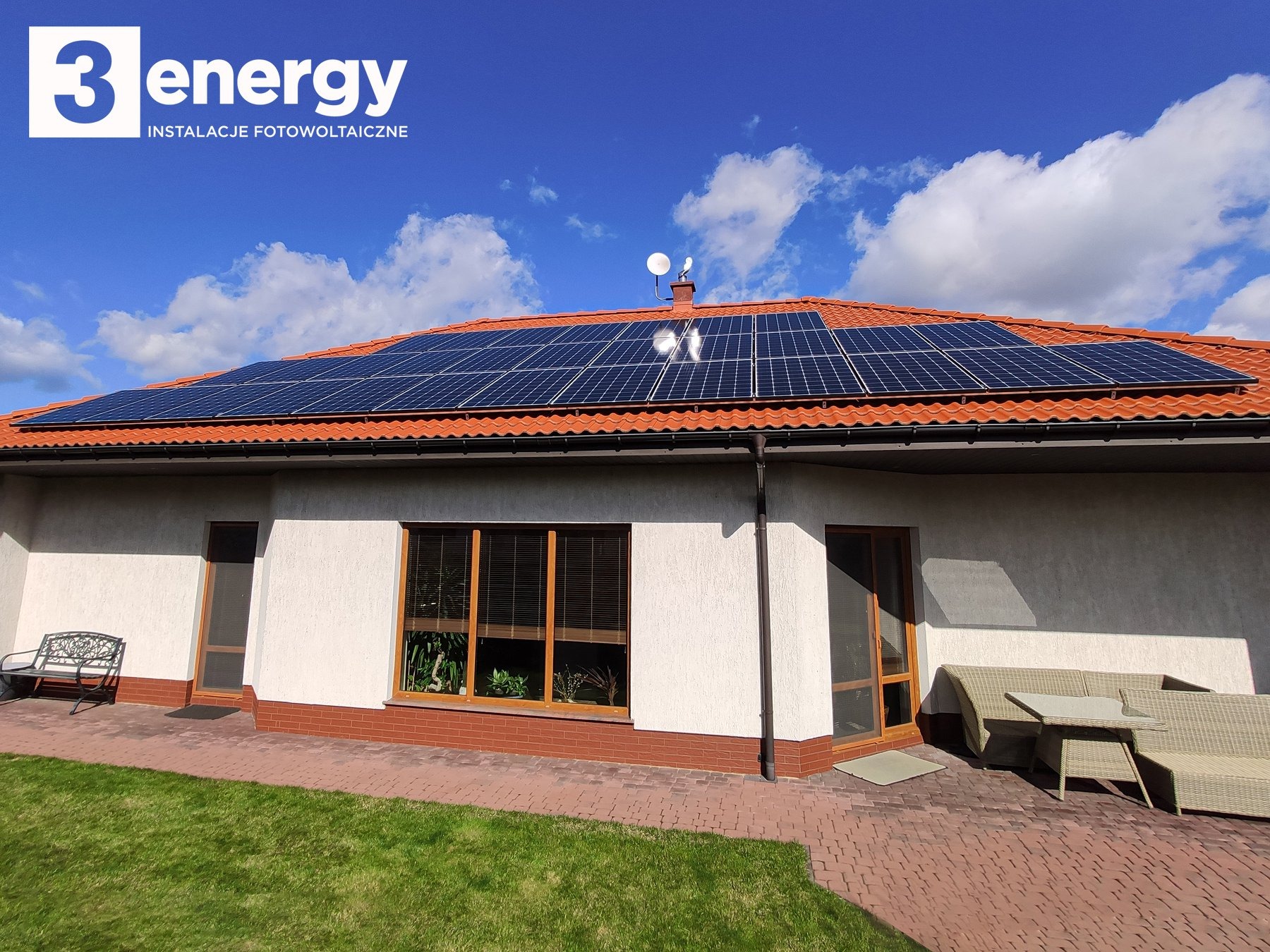Dom jednorodzinny z panelami słonecznymi na czerwonym dachu, widoczne logo firmy '3 energy' w lewym górnym rogu, błękitne niebo z chmurami w tle.
