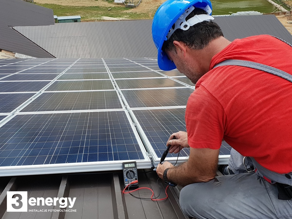 Instalator w niebieskim kasku mierzy napięcie na panelach słonecznych na dachu, używając multimetru; widoczne logo firmy instalacyjnej w rogu zdjęcia.