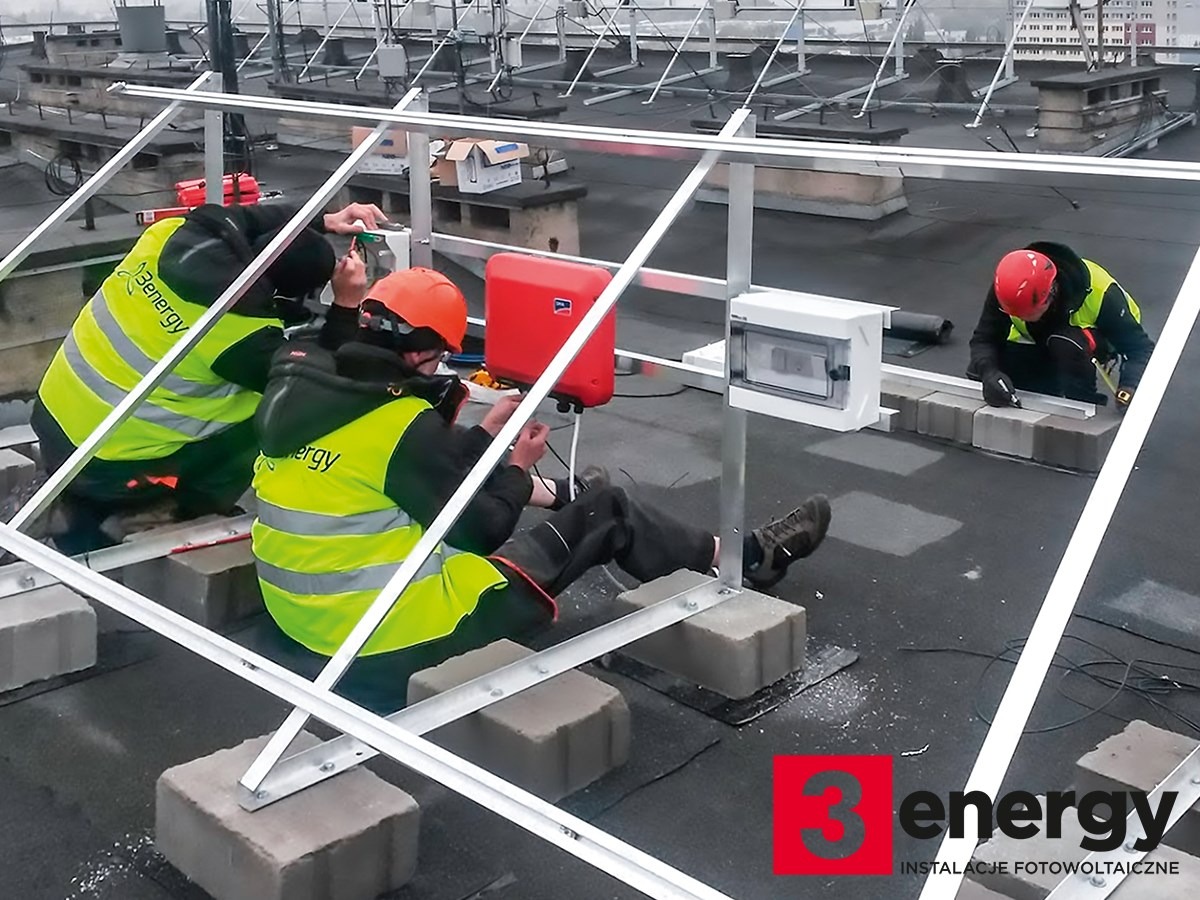 Montaż konstrukcji wsporczej paneli słonecznych na dachu płaskim; pracownicy w kamizelkach odblaskowych i kaskach, widoczna skrzynka elektryczna i falownik SMA, w tle inne konstrukcje