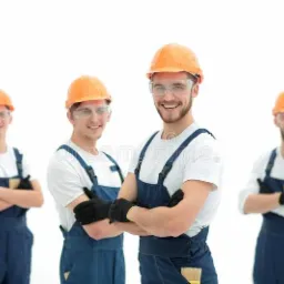 Grupa czterech uśmiechniętych pracowników w kombinezonach i kaskach ochronnych, z rękami założonymi, jeden z nich trzyma pędzel, na jasnym tle.