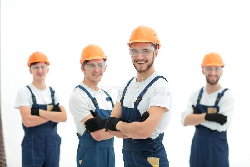 Grupa czterech uśmiechniętych pracowników w kombinezonach i kaskach ochronnych, z rękami założonymi, jeden z nich trzyma pędzel, na jasnym tle.