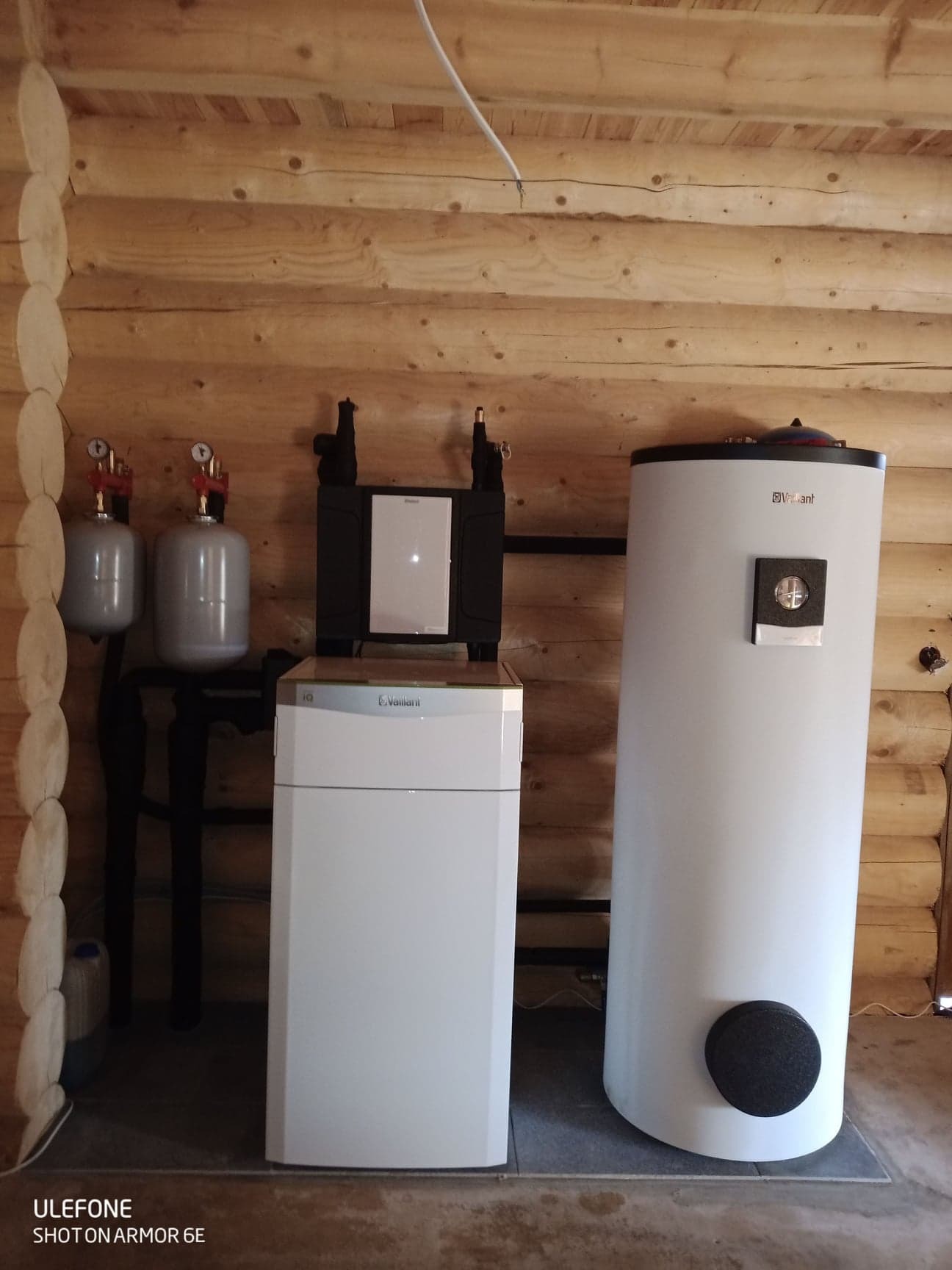 Instalacja grzewcza w drewnianym domu: biały zbiornik na wodę, jednostka sterująca Vaillant i dwa szare zbiorniki ciśnieniowe, połączone czarnymi rurami.