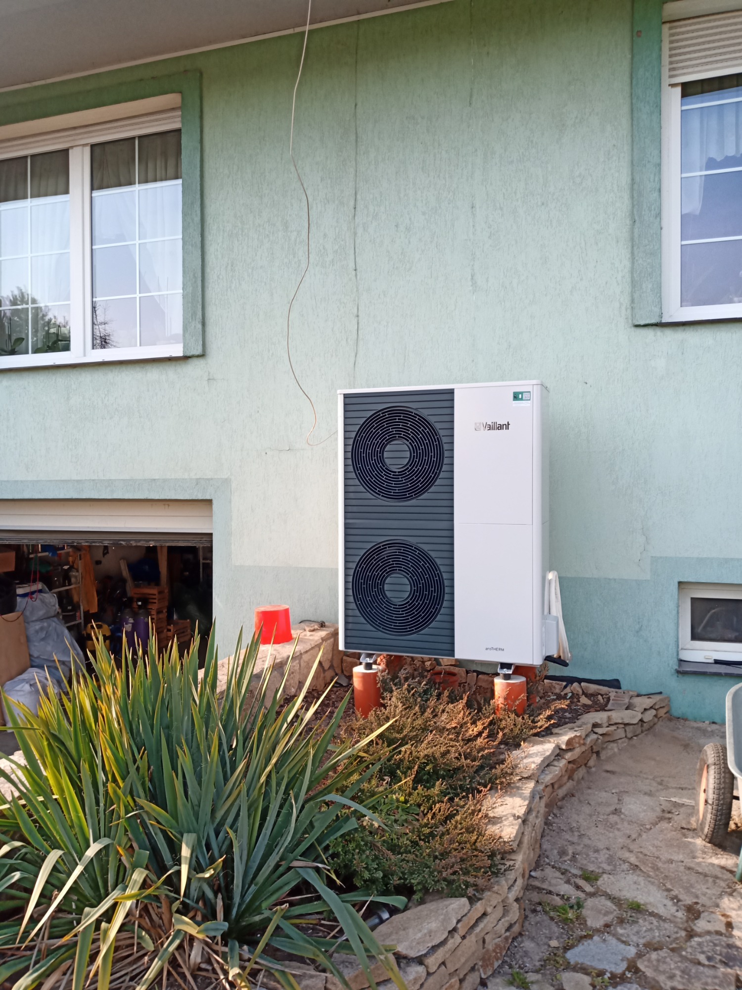 Biała pompa ciepła Vaillant AROTHERM zamontowana na zewnątrz budynku z zieloną elewacją, widoczne dwa wentylatory i elementy instalacji.