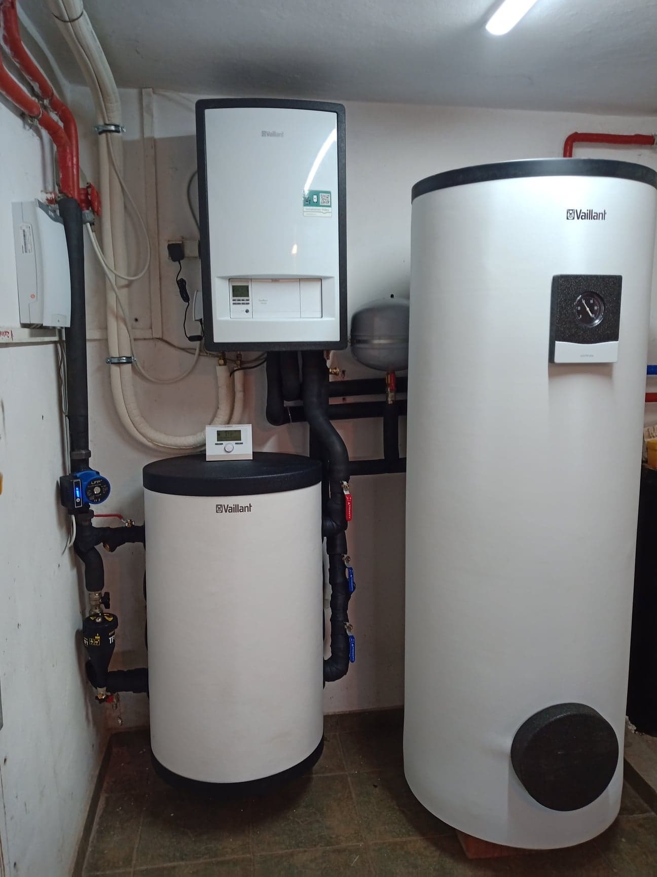 Zestaw urządzeń grzewczych Vaillant: wiszący piec, mniejszy i większy zasobnik ciepłej wody, rury instalacyjne w pomieszczeniu technicznym.