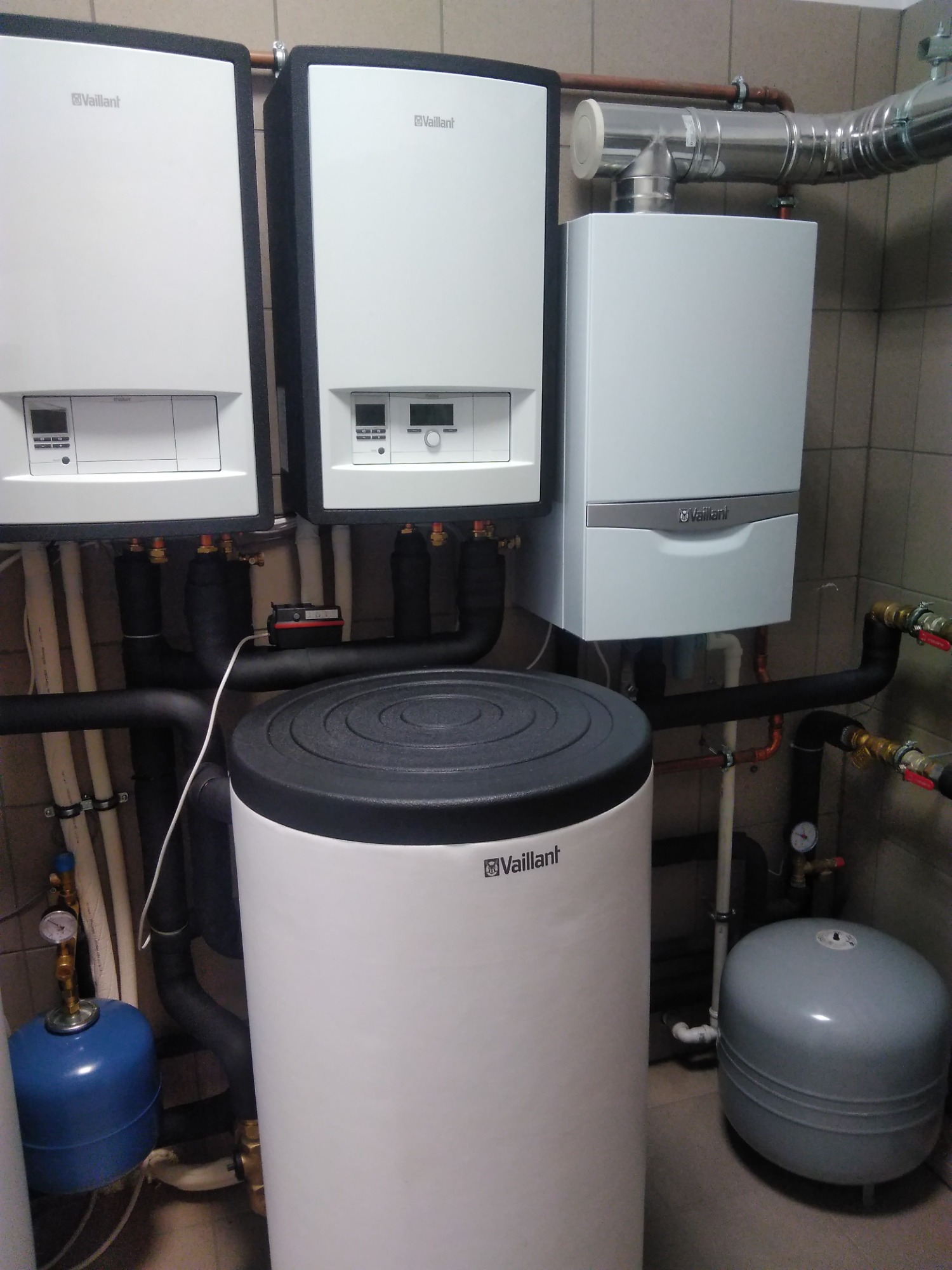 Dwa wiszące kotły grzewcze Vaillant obok siebie, z zasobnikiem ciepłej wody na dole i instalacją hydrauliczną w tle, na tle kafelków.