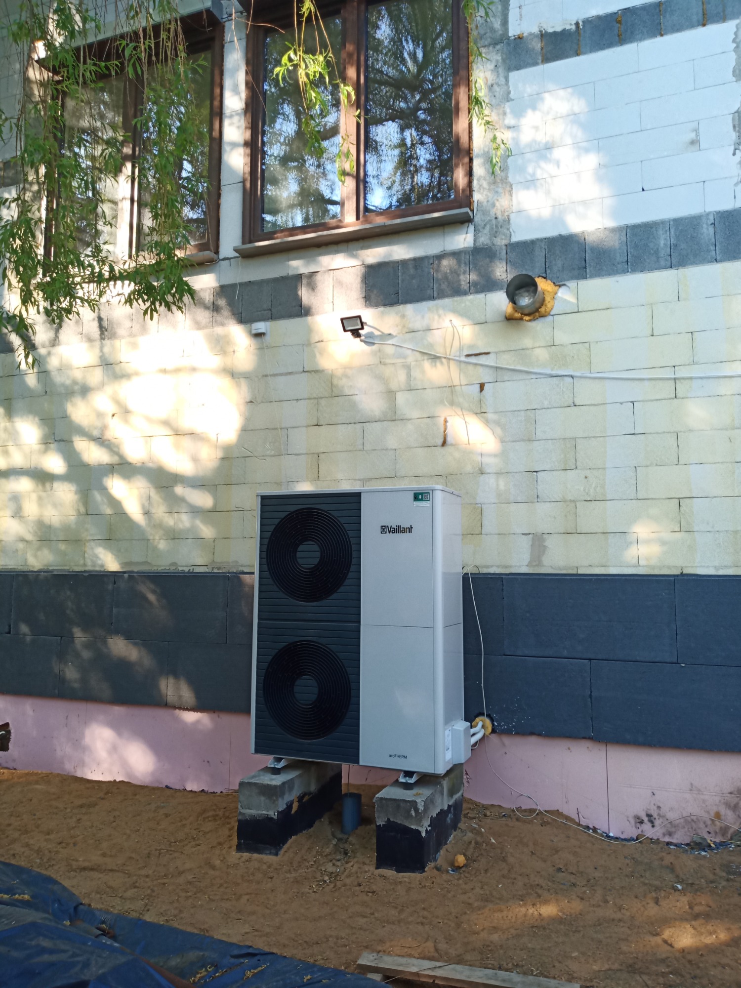 Biała pompa ciepła Vaillant, zamontowana na betonowych podstawach przy ścianie budynku z białego bloczka, w tle okno i gałęzie drzewa.