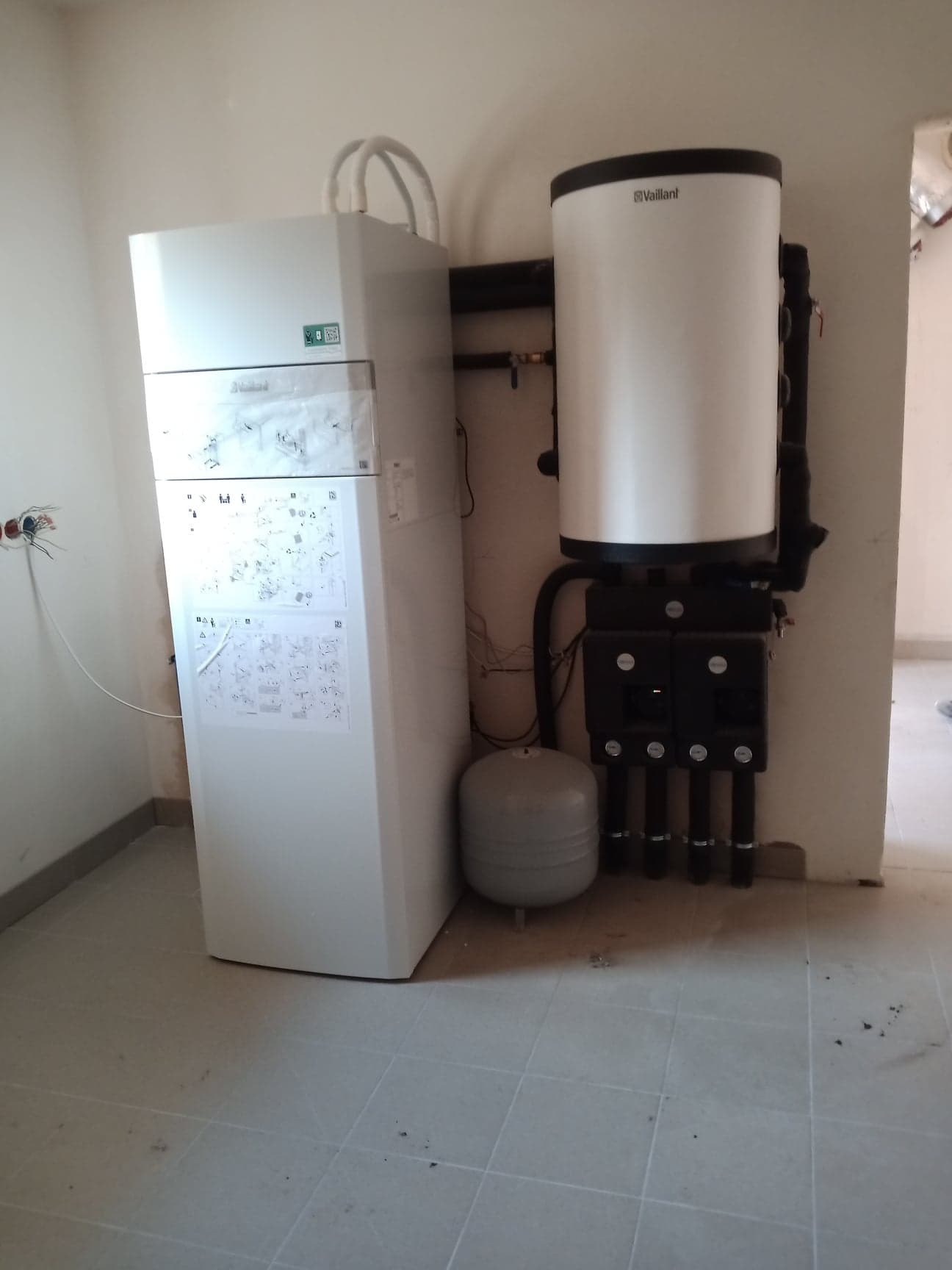 Nowa, biała instalacja grzewcza Vaillant z zbiornikiem i pompą ciepła w surowym pomieszczeniu z szarą płytką podłogową, widoczne rury i okablowanie.