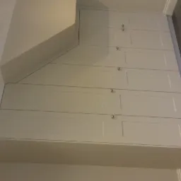 Biała szafa wnękowa z panelowymi drzwiami i srebrnymi gałkami, dopasowana do skosu sufitu, widoczne oświetlenie sufitowe.