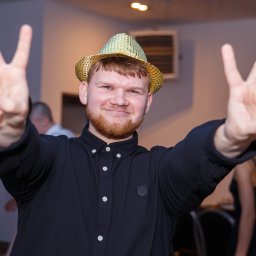 FOTO-WIDEO-KOŁO - Uśmiechnięty mężczyzna w czarnej koszuli i złotym kapeluszu pozuje, unosząc ręce w geście zwycięstwa na tle sali.