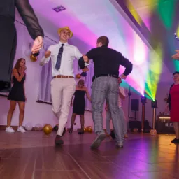 Roztańczona grupa osób na parkiecie podczas imprezy. W tle dekoracje z balonów i oświetlenie sceniczne. Mężczyzna w słomkowym kapeluszu w centrum kadru.