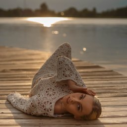FOTO-WIDEO-KOŁO - Kobieta w ażurowej sukience leży na drewnianym molo nad jeziorem w promieniach zachodzącego słońca. Ujęcie z niskiego kąta, podkreślające fakturę drewna i delikatność stroju.