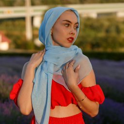 FOTO-WIDEO-KOŁO - Portret kobiety w czerwonym stroju i błękitnym hidżabie na tle pola lawendy. Ujęcie z dołu, delikatne światło podkreśla urodę modelki. W tle widoczna zieleń i fragment zabudowań.