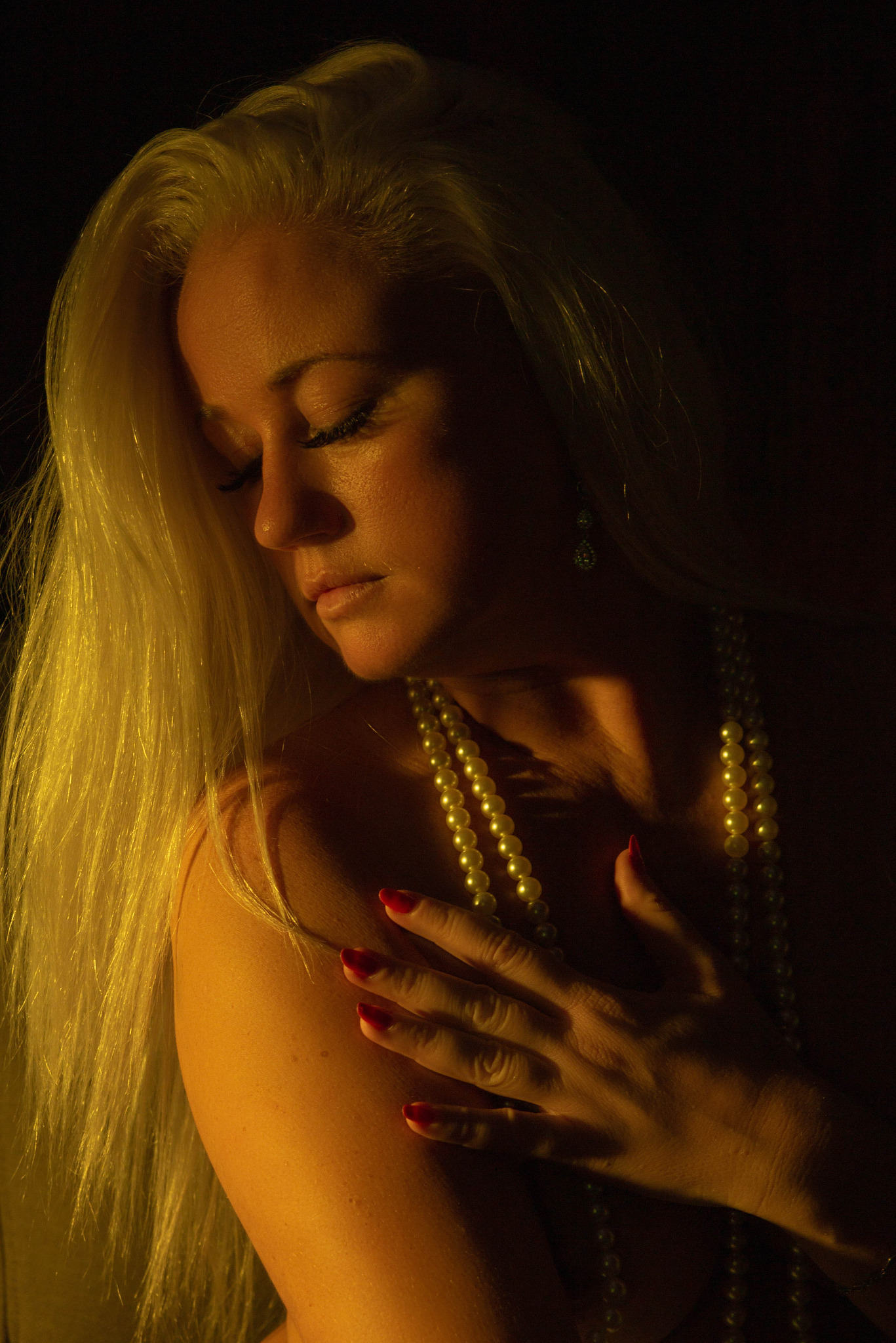 Portret kobiety z długimi blond włosami, spuszczonym wzrokiem, z perłami na szyi, oświetlonej ciepłym światłem, z czerwoną emalią na paznokciach.