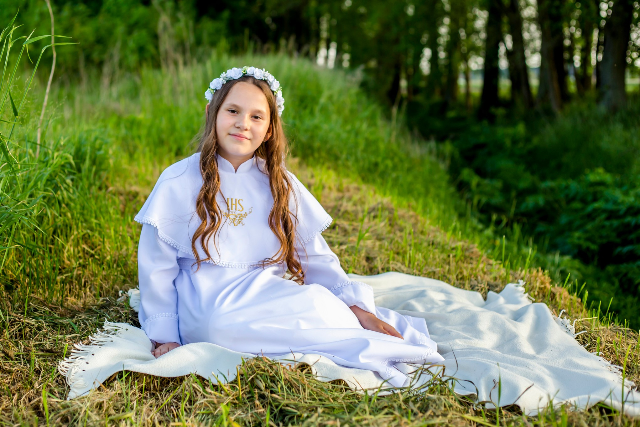 Dziewczynka w białej albie i wianku z białych kwiatów siedzi na kocu na trawie, w tle drzewa.