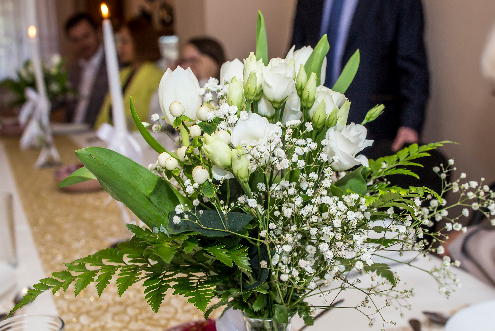 Dekoracyjny bukiet białych kwiatów i zieleni w szklanym wazonie na stole z złotym bieżnikiem, w tle osoby i zapalona świeca.