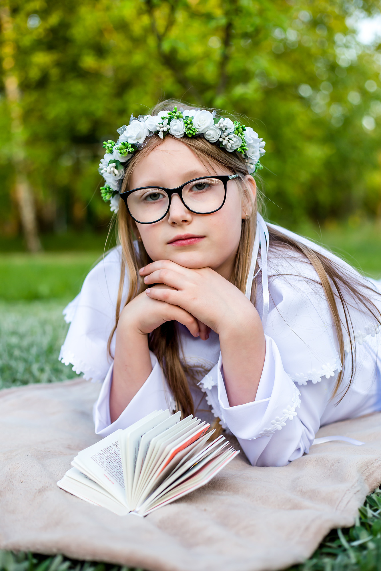 Dziewczynka w okularach i białej sukience komunijnej z wiankiem na głowie, leżąca na kocu z otwartą książką w dłoniach, na tle zieleni.