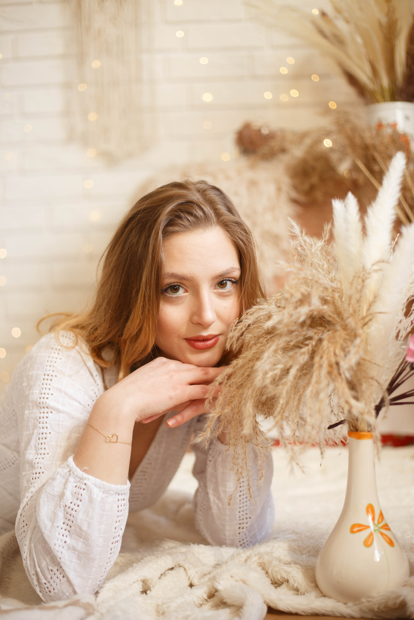 Portret kobiety w białej ażurowej bluzce, opierającej podbródek na dłoniach, z bransoletką w kształcie serca na nadgarstku, na tle puszystego dywanu i wazonów z trawą pampasową, oświetlony ciepłym...