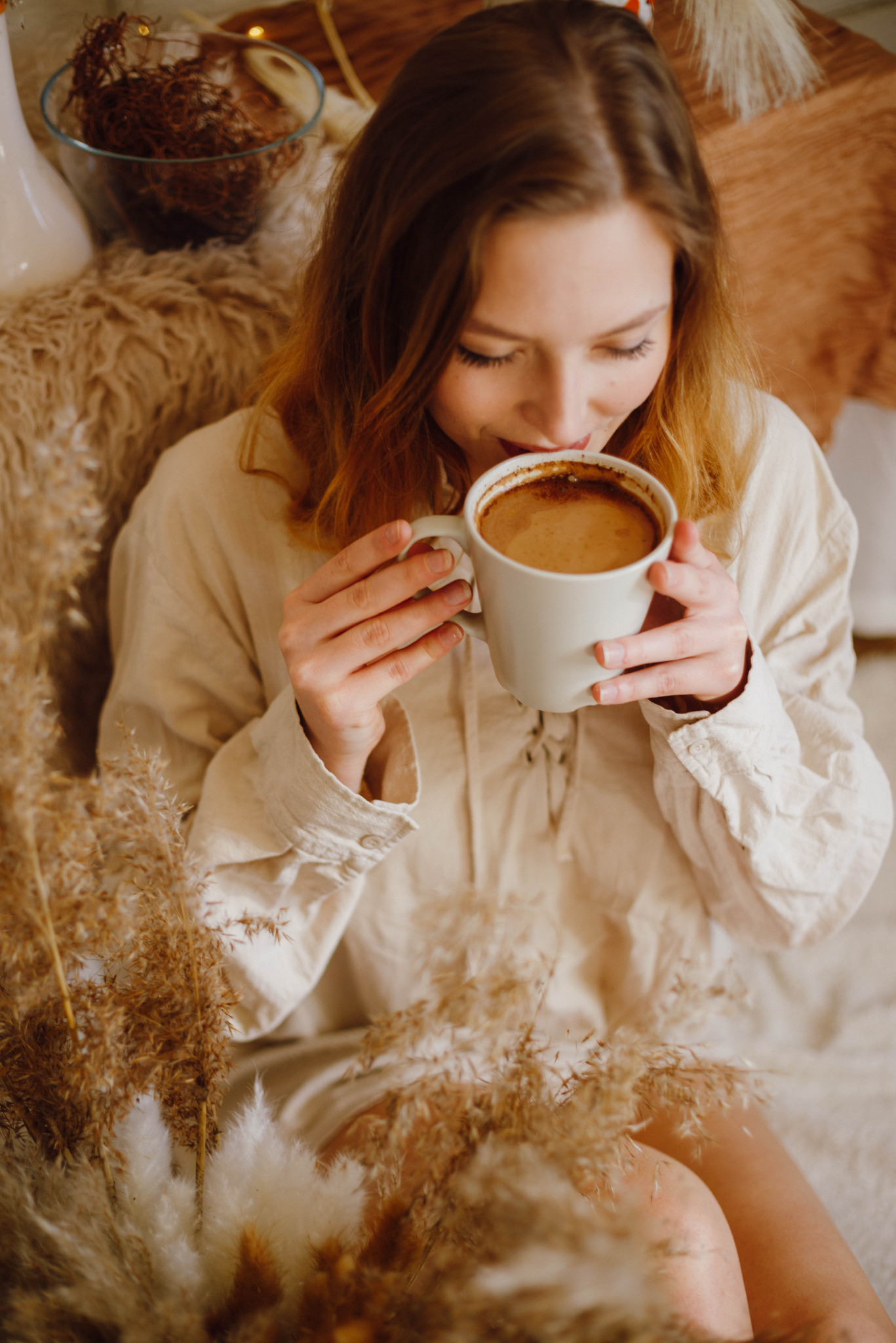 Młoda kobieta w beżowej koszuli pije kawę z białego kubka, otoczona dekoracyjnymi trawami w ciepłych odcieniach brązu i beżu, miękkie światło podkreśla naturalność sceny.