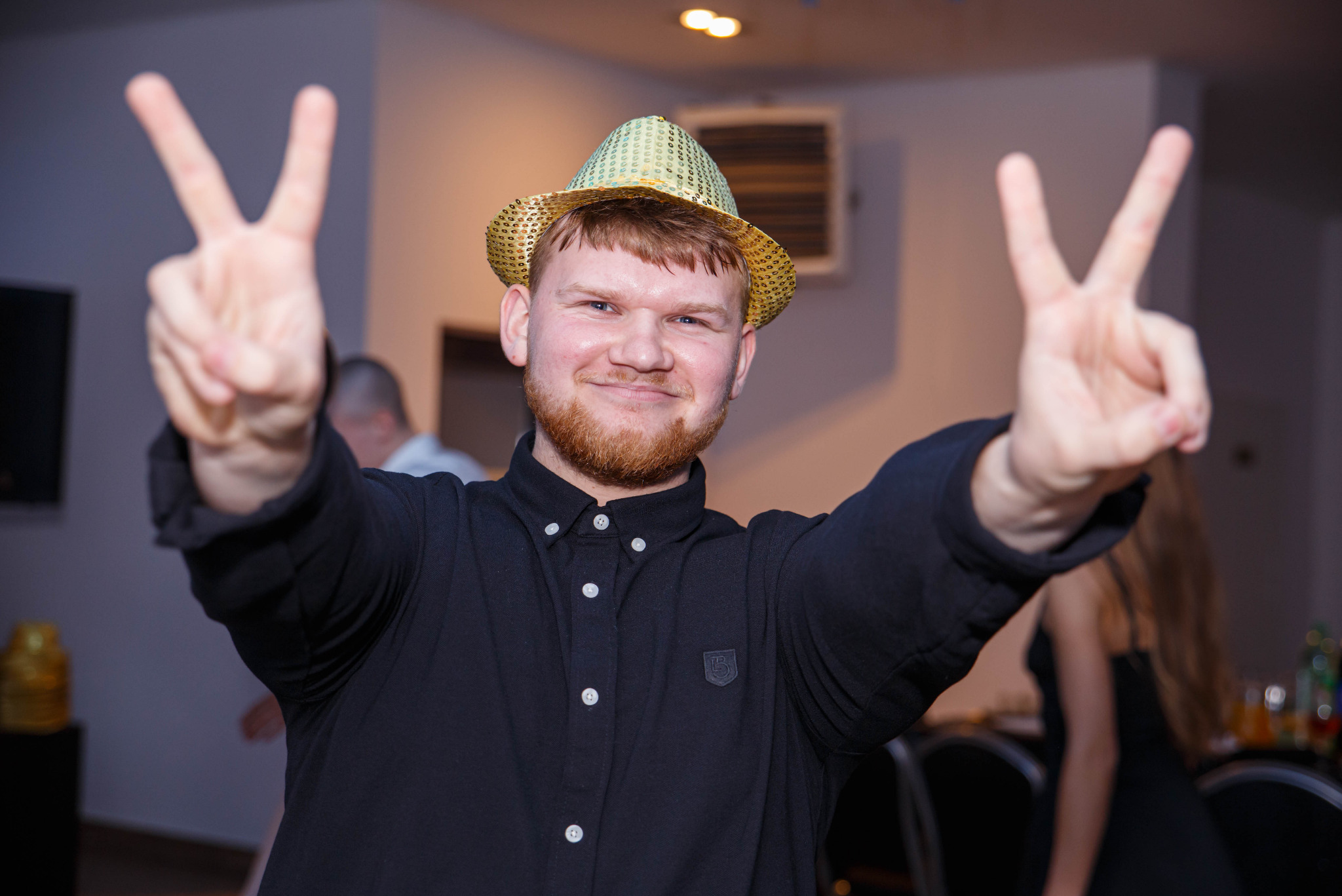 Uśmiechnięty mężczyzna w czarnej koszuli i złotym kapeluszu pozuje, unosząc ręce w geście zwycięstwa na tle sali.