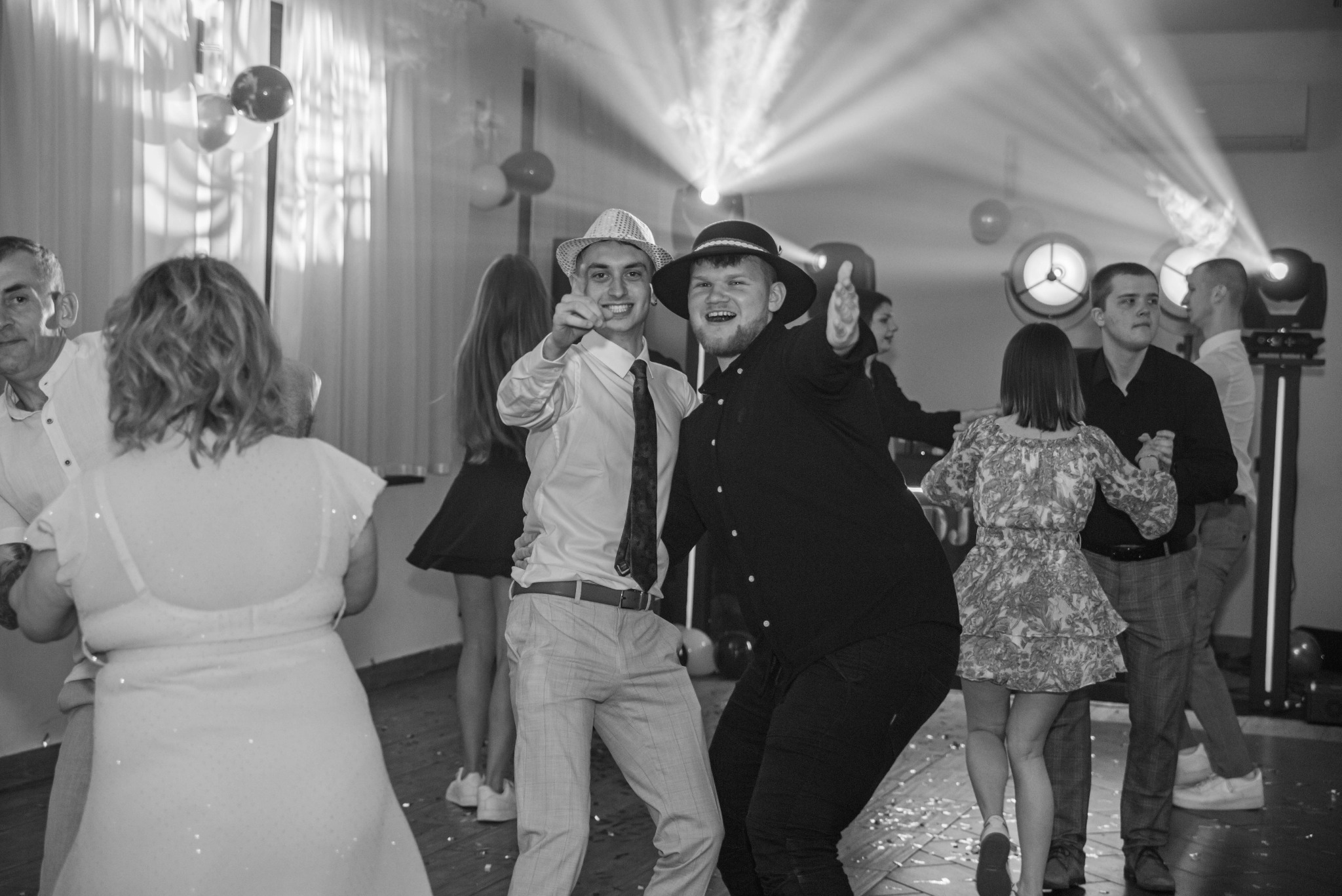 Czarno-białe ujęcie z imprezy: uśmiechnięci mężczyźni w kapeluszach tańczą w otoczeniu bawiących się gości, oświetleni światłami reflektorów.
