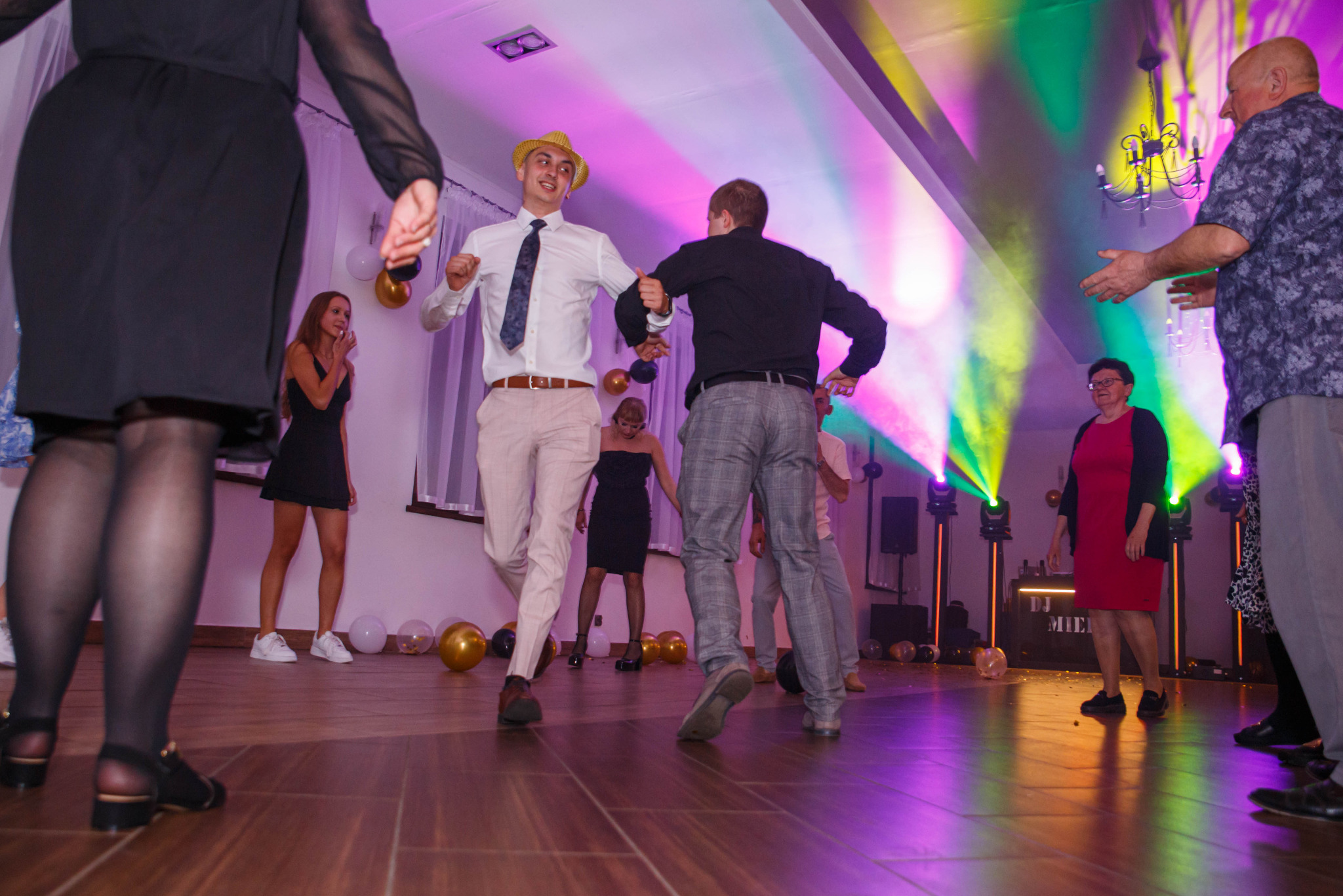 Roztańczona grupa osób na parkiecie podczas imprezy. W tle dekoracje z balonów i oświetlenie sceniczne. Mężczyzna w słomkowym kapeluszu w centrum kadru.