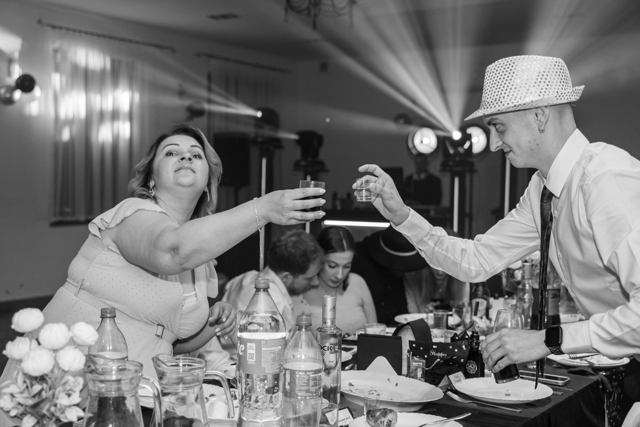 Czarno-białe zdjęcie z toastu na imprezie. Osoby podnoszą kieliszki przy stole zastawionym napojami i jedzeniem. Mężczyzna w kapeluszu i kobieta w sukience wznoszą toast.