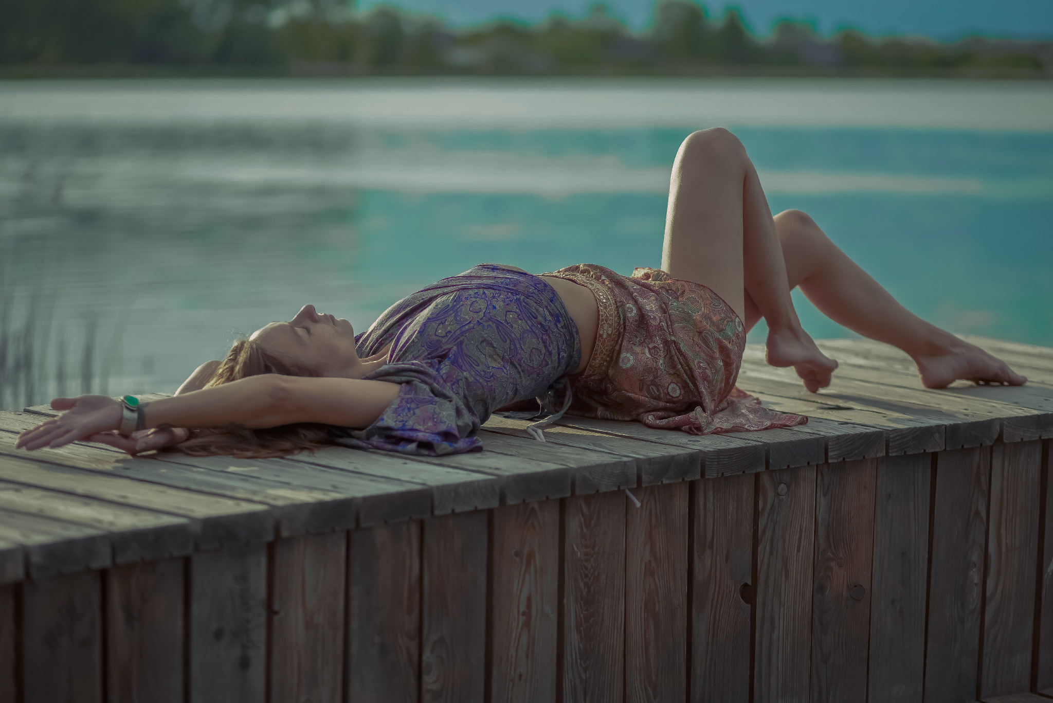 Kobieta relaksuje się, leżąc na drewnianym pomoście nad wodą. Ubrana w zwiewną sukienkę, z rozłożonymi rękami, patrzy w niebo. Spokojna sceneria i naturalne światło.