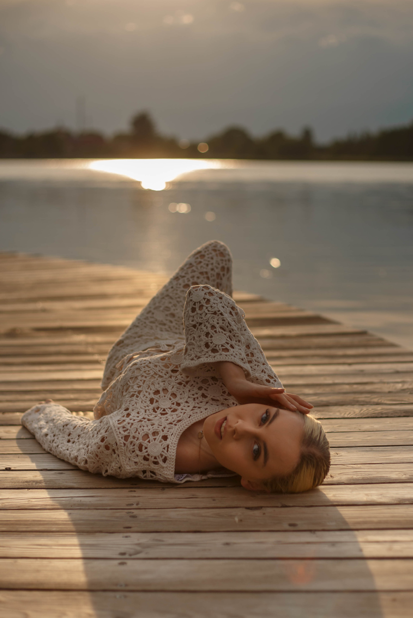 Kobieta w ażurowej sukience leży na drewnianym molo nad jeziorem w promieniach zachodzącego słońca. Ujęcie z niskiego kąta, podkreślające fakturę drewna i delikatność stroju.