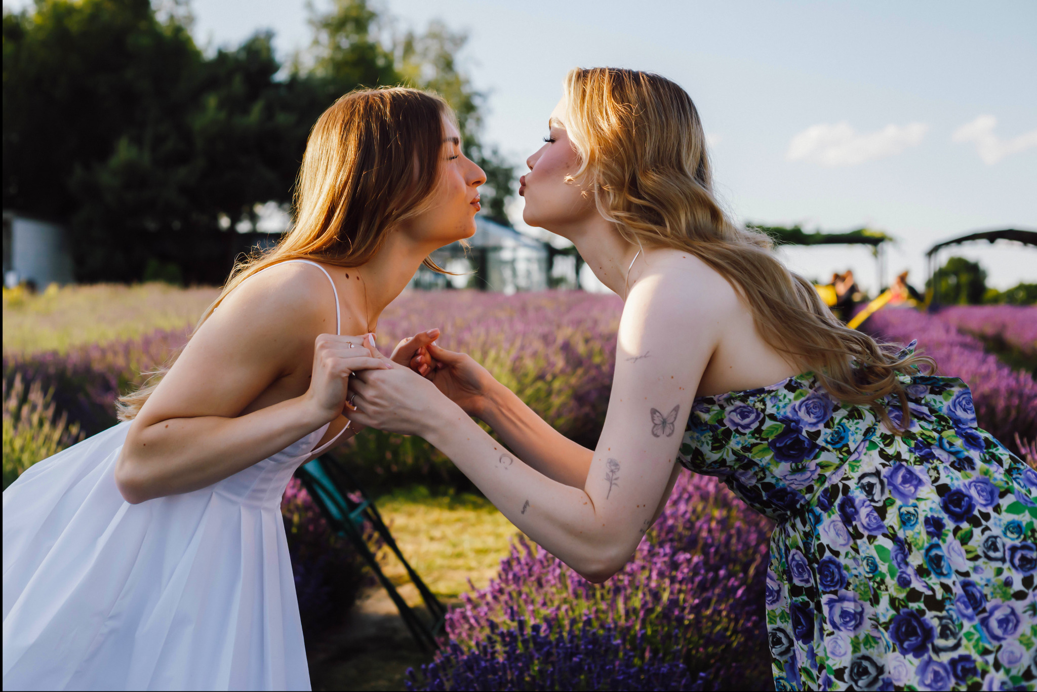 Dwie kobiety w kwiecistej sukience i białej sukni, stojące naprzeciw siebie na polu lawendy, z lekkim uśmiechem, w słoneczny dzień. Widoczne tatuaże na rękach.