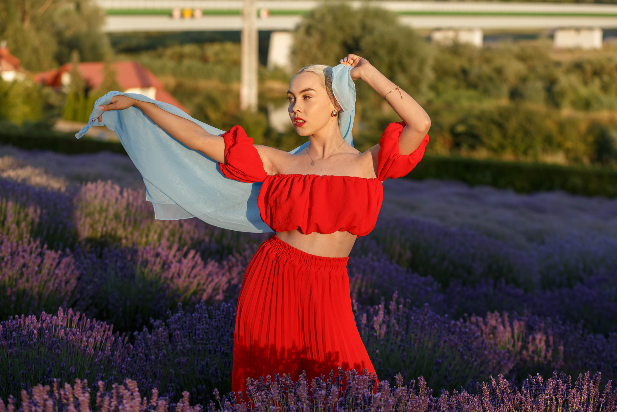 Blondwłosa kobieta w czerwonym stroju i błękitnym szalu pozuje na fioletowym polu lawendy, z widocznym mostem w tle, podczas sesji fotograficznej.